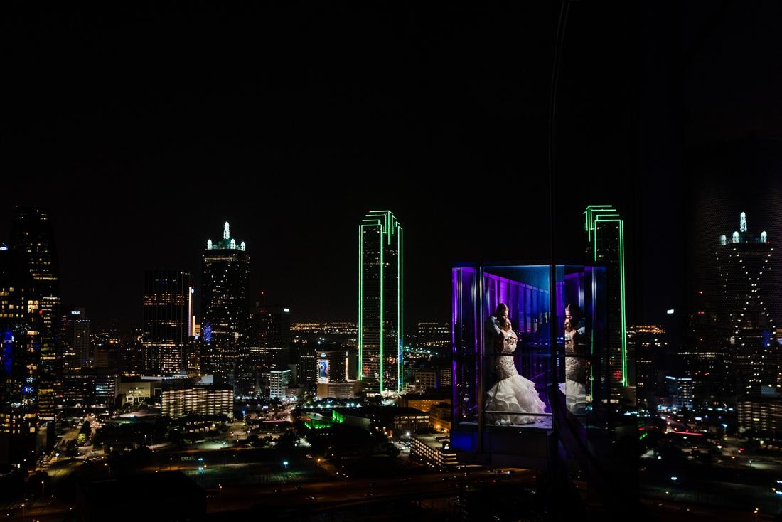 Couple embracing inside a lit glass elevator above the Dallas skyline at night